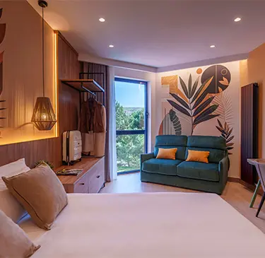 Habitación moderna con cama, sofá, mural colorido y ventana con vista al exterior.