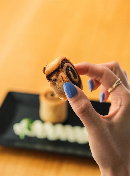 Mano sosteniendo un pequeño rollo de canela con manicura azul, bandeja negra al fondo.