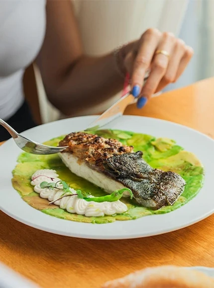 Persona comiendo pescado con guarnición sobre salsa verde en un plato blanco.