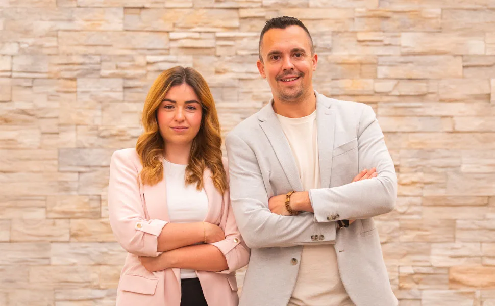 Dos personas sonrientes, de pie, con fondo de pared de piedra.