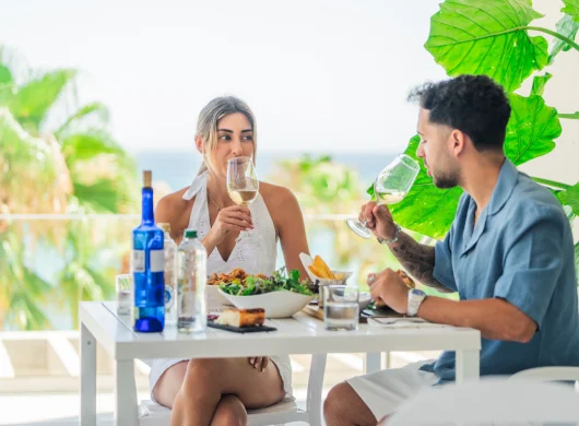 Pareja disfrutando de vino y ensaladas en una terraza soleada.