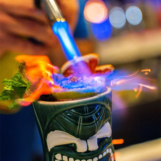 Cóctel en vaso tiki con llamas azules y naranjas, adornado con menta y limón.