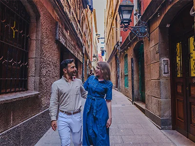 Una pareja sonriente camina por una calle estrecha y adoquinada, rodeada de edificios antiguos.