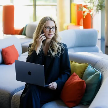 Mujer con gafas usando laptop y teléfono en un sofá, rodeada de cojines coloridos.