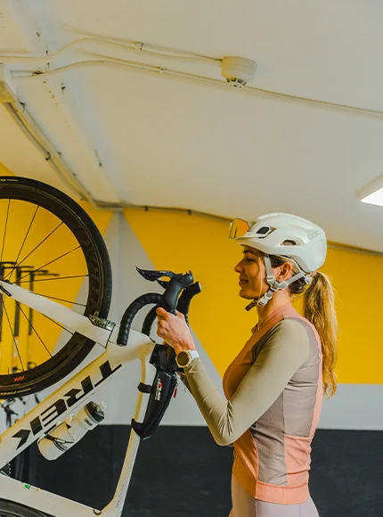 Mujer con casco ajustando una bicicleta blanca en un espacio interior con paredes amarillas.