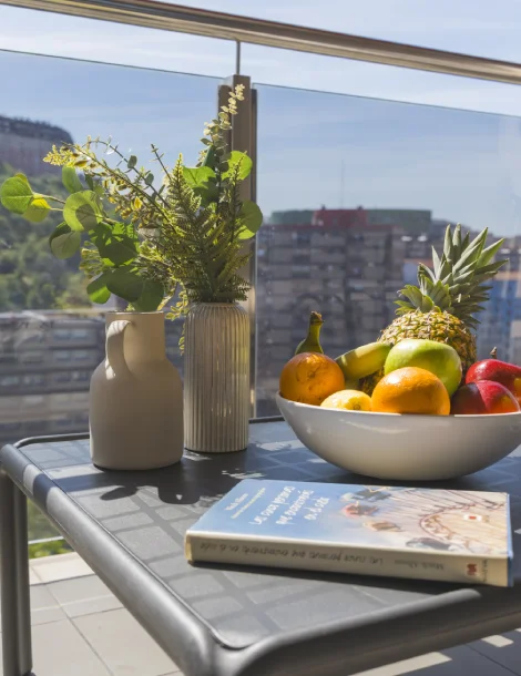 Mesa con frutas, jarrones y un libro en un balcón con vista a la ciudad.