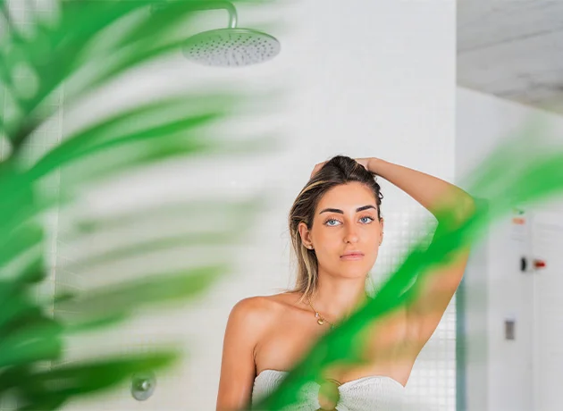 Mujer en una ducha con hojas verdes en primer plano, mirando relajada hacia adelante.