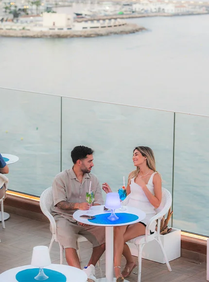 Pareja sentada en una terraza frente al mar, disfrutando de bebidas y conversando.