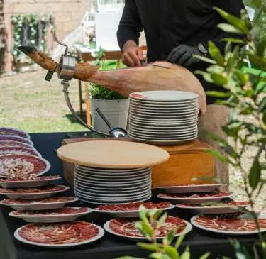 Persona cortando jamón con platos preparados al aire libre.