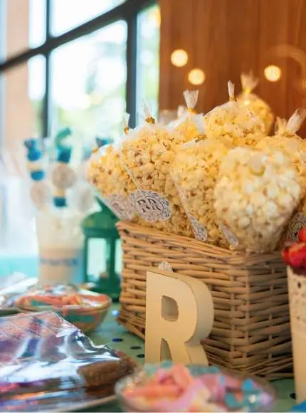 Cesta con bolsas de palomitas, letras decorativas y dulces en una mesa de fiesta.