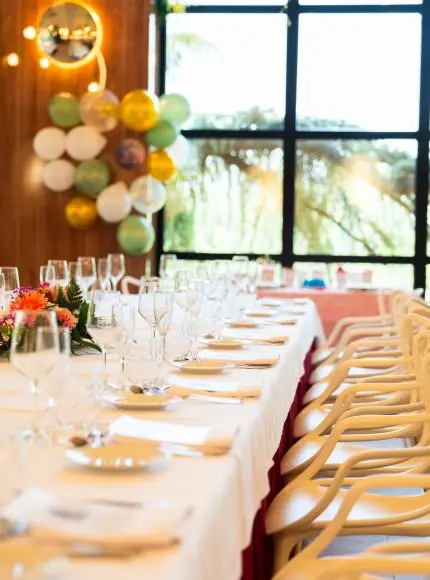 Mesa elegante con copas de vino y vajilla, decorada con flores y globos, junto a una ventana.
