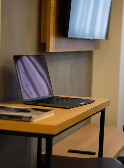 Portátil abierto sobre una mesa de madera junto a una pared y un televisor montado.