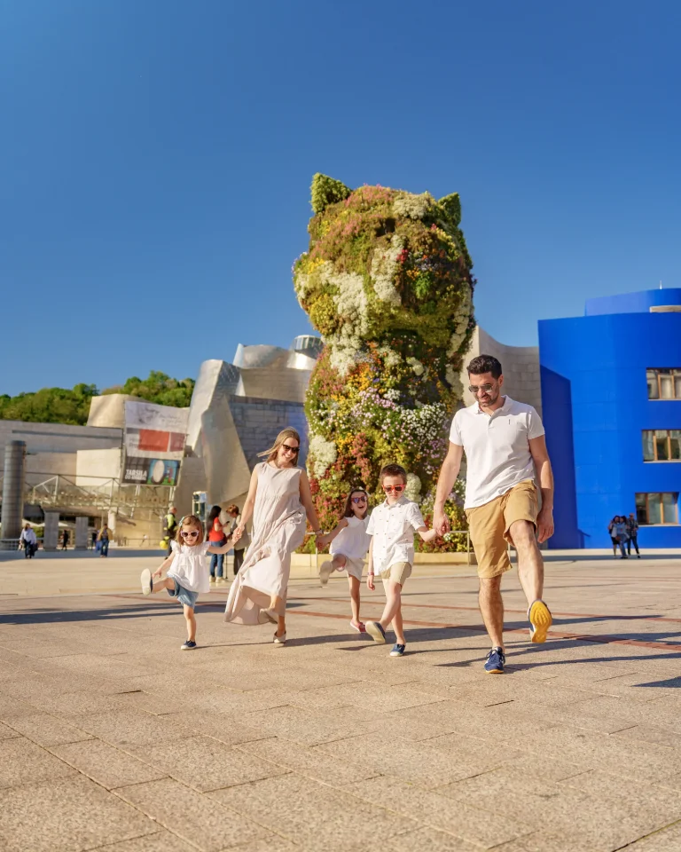Familia caminando frente a una gran escultura floral de cachorro bajo un cielo despejado.