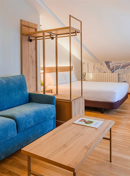 Habitación moderna con cama, sofá azul y mesa de madera en ambiente luminoso.