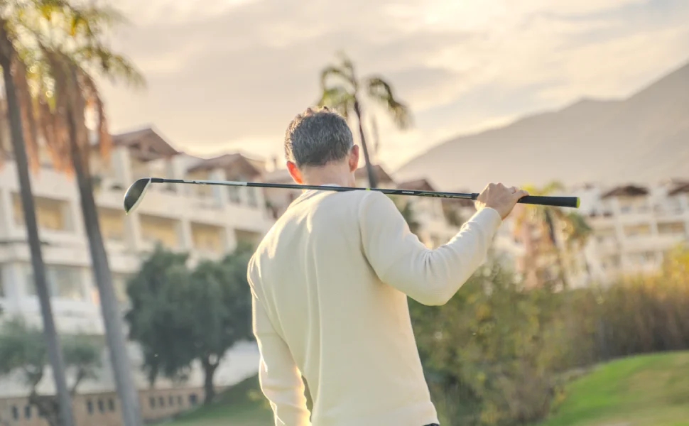 Hombre cargando un palo de golf en un campo, con palmeras y edificios al fondo.