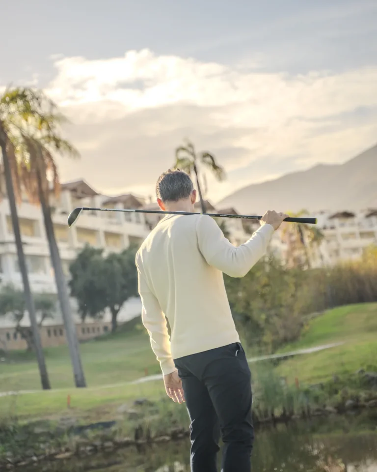 Persona de espaldas con un palo de golf en el hombro, con paisaje de árboles y edificios al fondo.