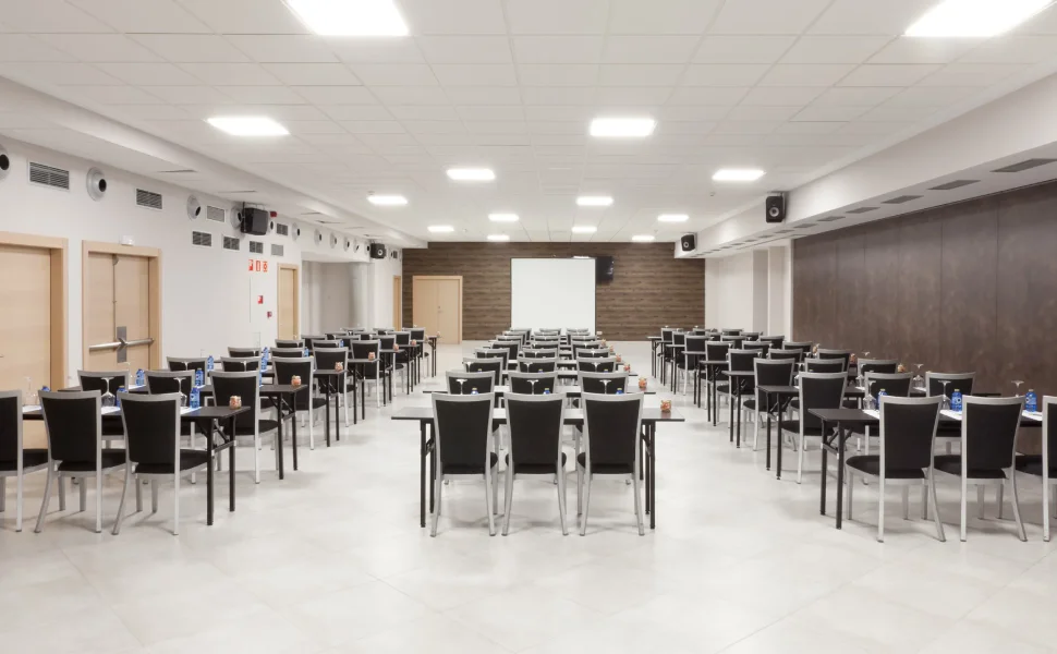 Sala de conferencias vacía con mesas y sillas, frente a una pantalla blanca.