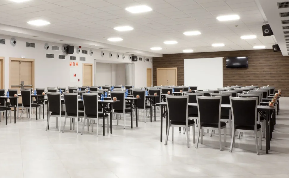 Sala de conferencias vacía con sillas, mesas y proyector blanco en el fondo.