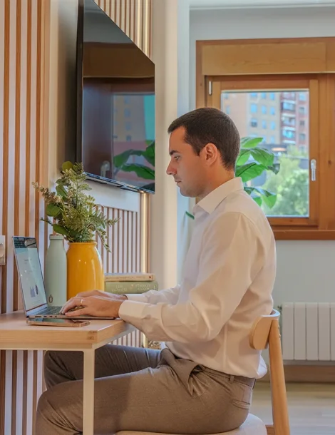 Hombre trabajando en laptop cerca de ventana con plantas.
