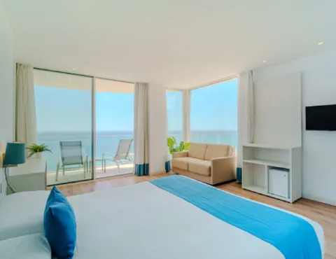 Habitación luminosa con cama, sofá, balcón con vista al mar y decoración en tonos blancos y azules.