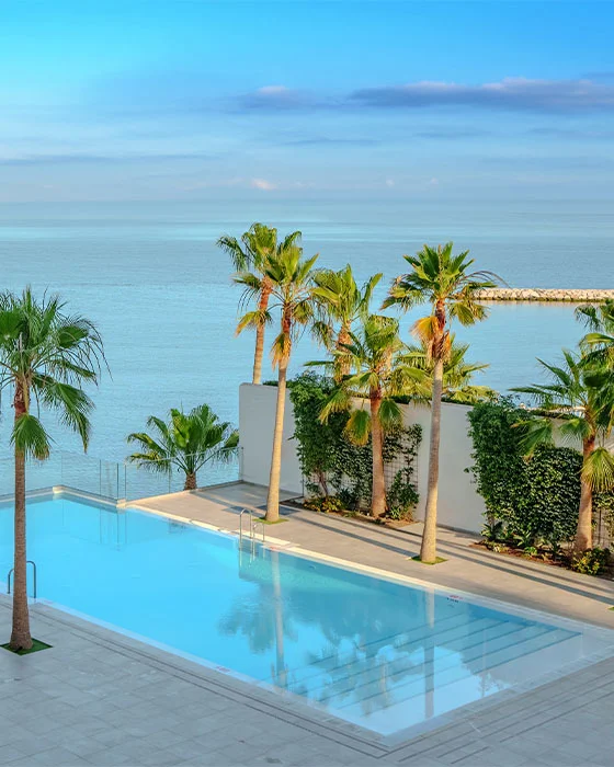 Piscina junto a la playa con palmeras y cielo azul brillante.
