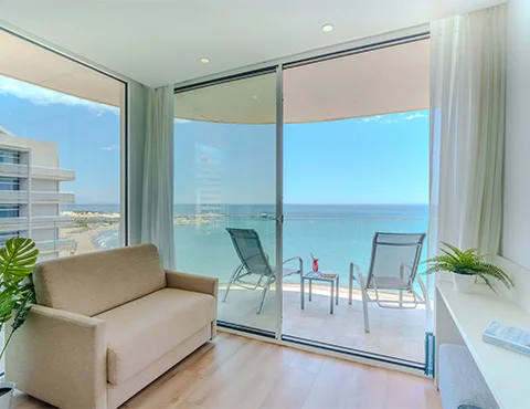 Sala moderna con vista al mar desde un balcón acristalado, sofá y sillas exteriores.