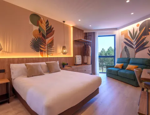 Habitación moderna con cama, sofá, decorada con hojas y luz cálida, vista al exterior.