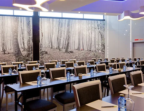 Sala de conferencias con mesas, sillas y botellas de agua, fondo de bosque en una pared.