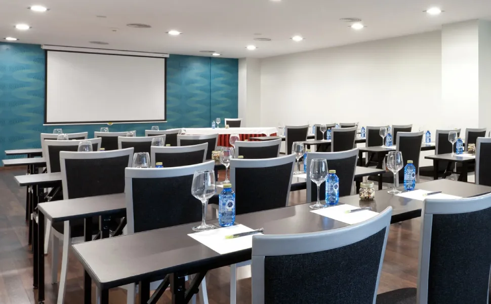 Sala de conferencias con mesas dispuestas, botellas de agua y copas vacías.