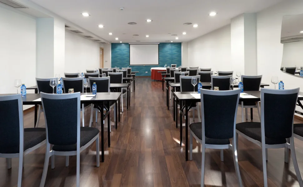 Sala de conferencias lista con mesas, sillas, pantalla y botellas de agua.