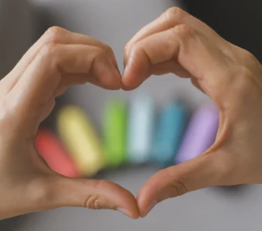 Manos formando un corazón con tizas de colores desenfocadas al fondo.