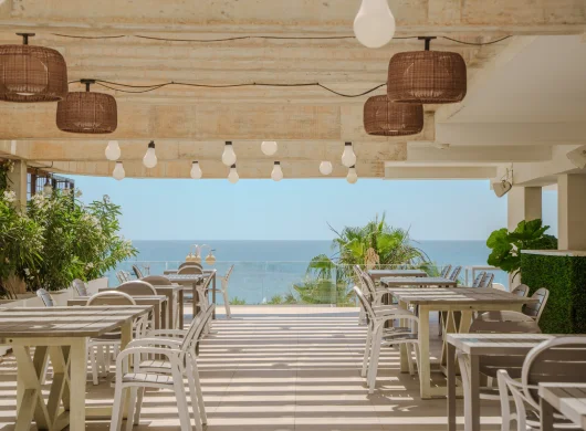 Terraza con mesas y vista al mar, decorada con lámparas colgantes y plantas.