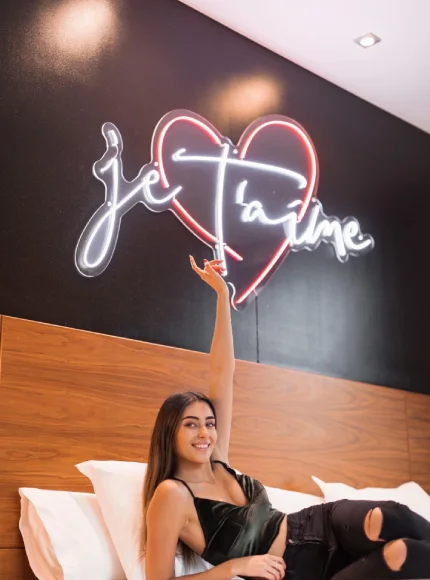 Mujer sonriendo bajo letrero de neón "je t'aime" con corazón.