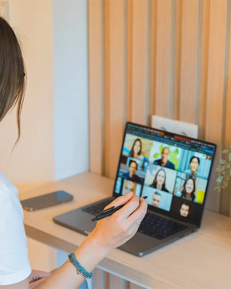 Persona en videollamada en laptop, sujetando un lápiz, con un teléfono al lado.