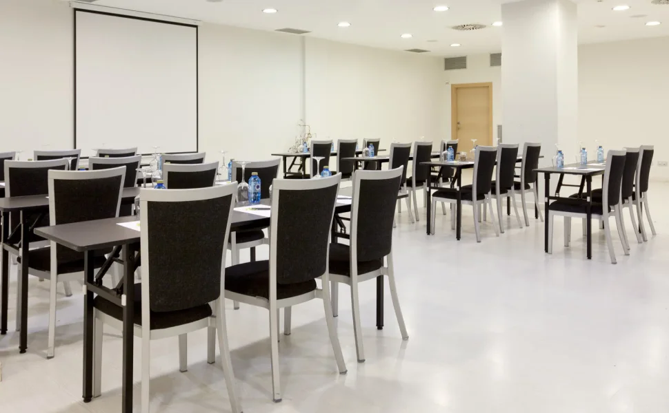 Sala de conferencias vacía con sillas, mesas y pantalla de proyección.