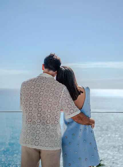 Pareja abrazada mirando el mar desde un balcón, en un día soleado.