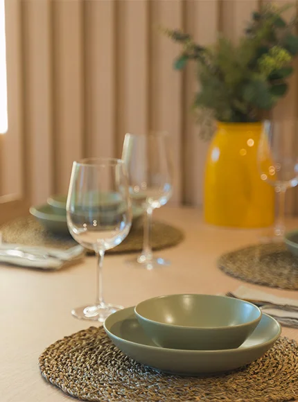Mesa elegante con platos verdes y copas, jarra amarilla al fondo.