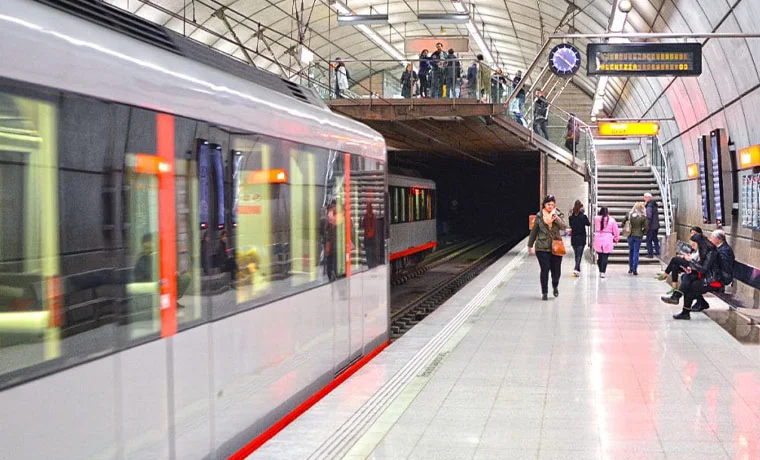 Estación de metro con un tren en movimiento y varias personas en el andén.