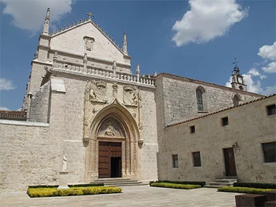 Iglesia de piedra con puerta gótica, esculturas y una plaza con cielo azul y nubes.