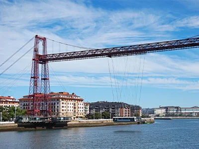 Puente colgante rojo cruzando un río, con edificios al fondo bajo un cielo azul.