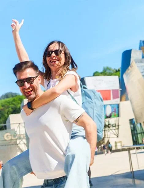 Pareja sonriente en la calle, ella sobre su espalda con el brazo al aire.