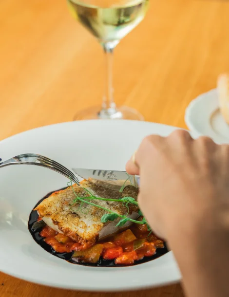 Persona cortando pescado en plato, acompañado de vino blanco.