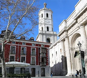 Torre y edificio histórico en una plaza con cielo azul y árboles sin hojas.