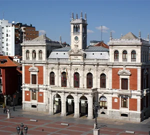 Edificio histórico con torre y arco en una plaza urbana, rodeado de edificios modernos.