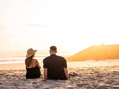 Pareja sentada en la playa durante la puesta de sol, mirando al mar.