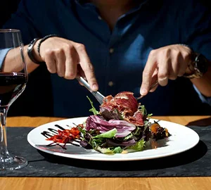 Persona en restaurante cortando ensalada en un plato, con copa de vino al lado.