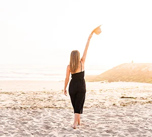 Persona de pie en la playa al atardecer, levantando un sombrero hacia el cielo.