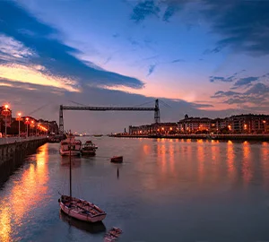 Atardecer en un río con un puente colgante, barcos anclados y luces de la ciudad reflejadas en el agua.