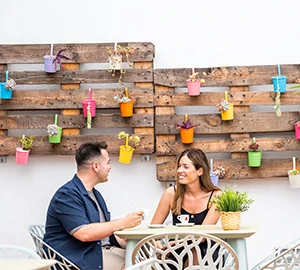 Dos personas conversando en un café al aire libre, con decoración de macetas coloridas en la pared.