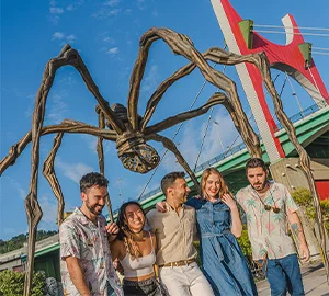 Grupo de personas posando frente a una gran escultura de araña y un puente rojo.